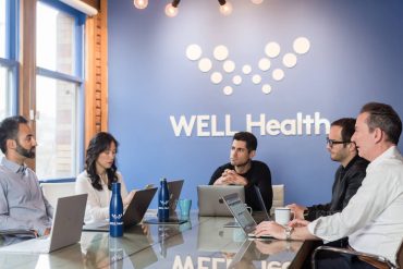 a group of five people is meeting in a board room with the logo of the Well Health company on the wall