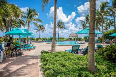 The photo shows a resort with a big swimming pool in the middle surrounded by blue umbrellas and palm trees during the day.
