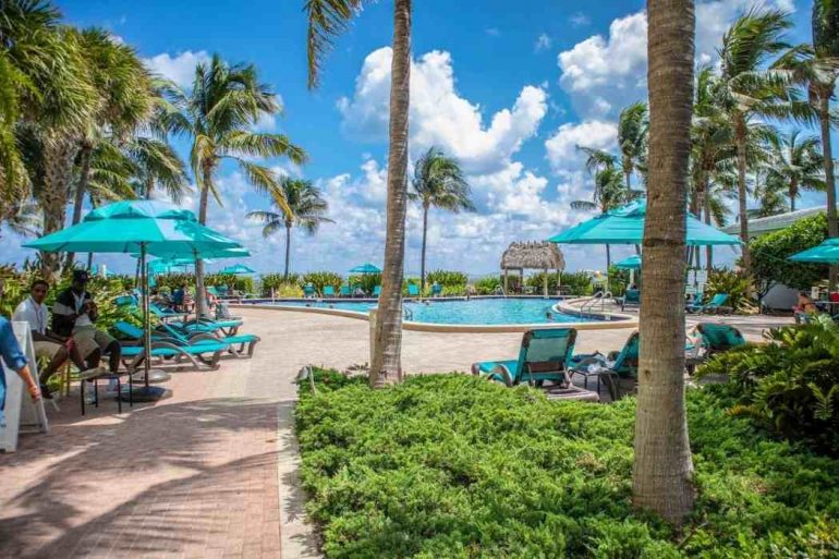 The photo shows a resort with a big swimming pool in the middle surrounded by blue umbrellas and palm trees during the day.