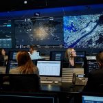 An image featuring the inside of IBM Canada's facility. Multiple people are sitting down in their designated spots with a monitor screen in front of them. All of the employees are facing two large monitors displaying a map and analytics.