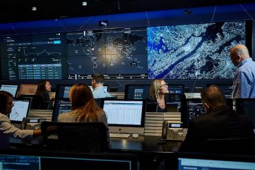 An image featuring the inside of IBM Canada's facility. Multiple people are sitting down in their designated spots with a monitor screen in front of them. All of the employees are facing two large monitors displaying a map and analytics.