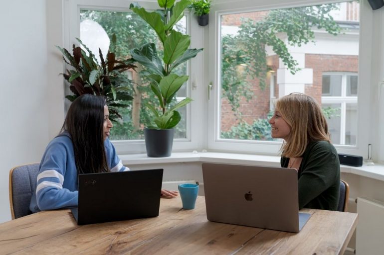 Two people facing each other while they sit down by a wooden table with both their laptops open.