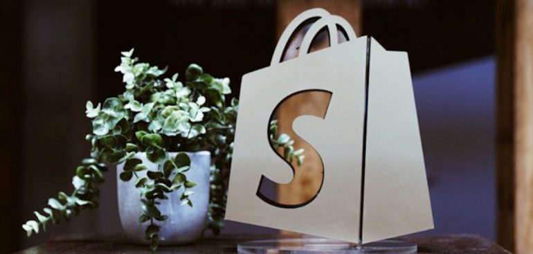 Shopify logo bag beside a green plant.
