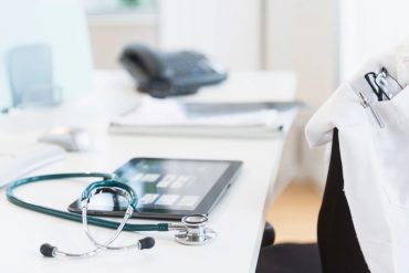 Stethoscope lying on an ipad which is on a desk. On the chair of the desk there is a white lab coat.
