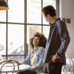 two people talk at a coffee shop. A woman is seated and an employee takes her order