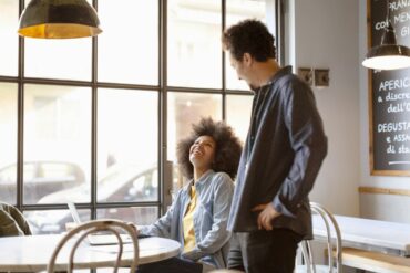 two people talk at a coffee shop. A woman is seated and an employee takes her order