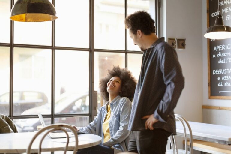 two people talk at a coffee shop. A woman is seated and an employee takes her order