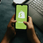 A pair of hands holding a mobile phone which has the Shopify logo on it