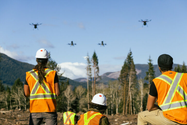three workers in visibility vests observe as drones take off over burn landscape with mountains and forest in the background