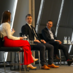 One woman and two men speak on a panel at CityAge. The woman is facing the two men in suits. She appears to be moderating