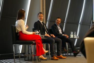 One woman and two men speak on a panel at CityAge. The woman is facing the two men in suits. She appears to be moderating