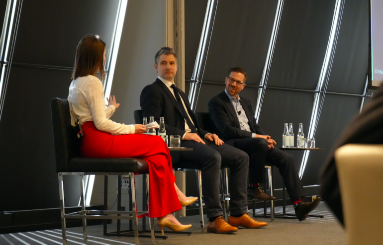 One woman and two men speak on a panel at CityAge. The woman is facing the two men in suits. She appears to be moderating
