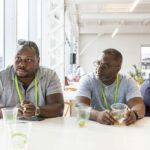 Two men wearing lanyards sit at a table with plastic cups in front of them at a past startupfest event