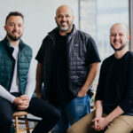 The three founders of OneVest pose for a portrait in an office with floor to ceiling windows.