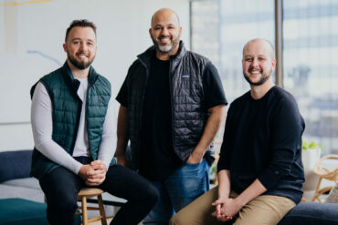 The three founders of OneVest pose for a portrait in an office with floor to ceiling windows.