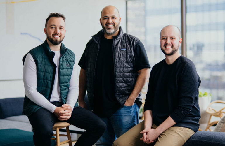 The three founders of OneVest pose for a portrait in an office with floor to ceiling windows.