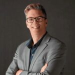 portrait of Darrell Petras on a plain grey background. he wears a suit and black eyeglasses.