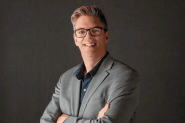 portrait of Darrell Petras on a plain grey background. he wears a suit and black eyeglasses.