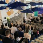 people sit in edmonton unlimited's event space watching a female speaker. cool lighting fixtures that look like umbrellas are overhead. there's a steeply tiered seating area and about 60-70 people in attendance