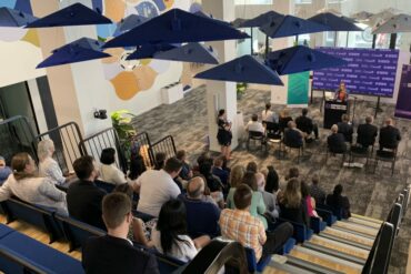people sit in edmonton unlimited's event space watching a female speaker. cool lighting fixtures that look like umbrellas are overhead. there's a steeply tiered seating area and about 60-70 people in attendance