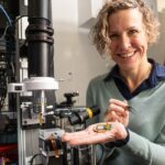 Photonic co-founder Stephanie Simmons holding a chip in her raised hand. She is smiling and appears to be in some kind of workshop.