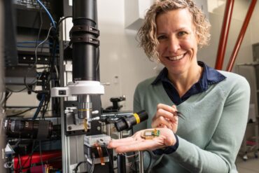 Photonic co-founder Stephanie Simmons holding a chip in her raised hand. She is smiling and appears to be in some kind of workshop.