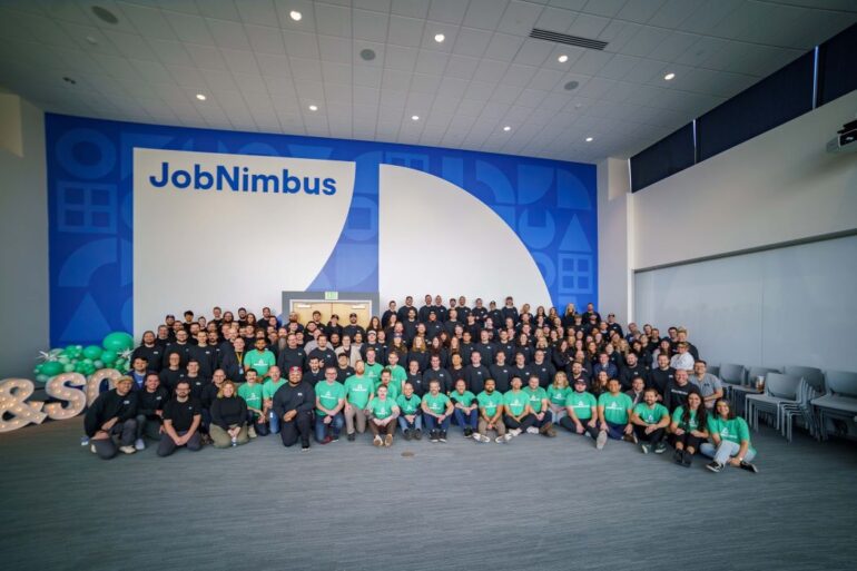 A group photo of the JobNimbus and SumoQuote employees in front of a wall with JobNimbus branding. Some are wearing SumoQuote-style mint green shirts, while many are wearing dark blue.
