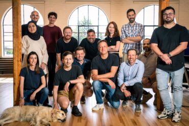 14 people, of whom three are women, pose for the camera in a staff portrait in an empty room with large arched windows and wooden floors. a golden retreiver sleeps at their feet.