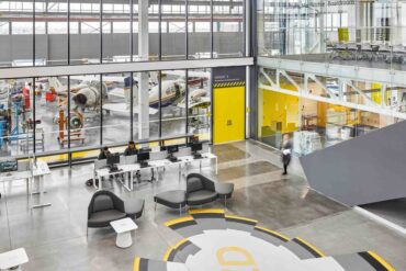 A view from above a bright, lab-style room with large windows looking into a factory floor containing several small airplanes