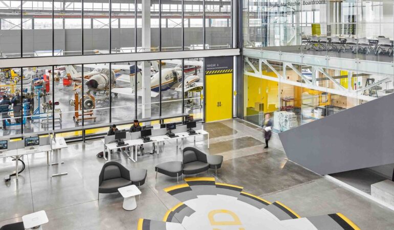 A view from above a bright, lab-style room with large windows looking into a factory floor containing several small airplanes
