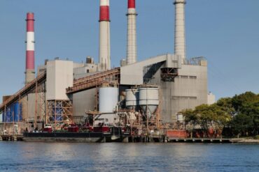 A refinery with large smokestacks on the banks of an unknown body of water
