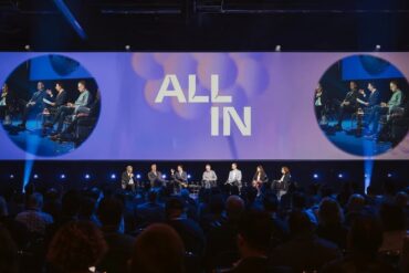 Seven people sit in a row onstage in front of a large crowd