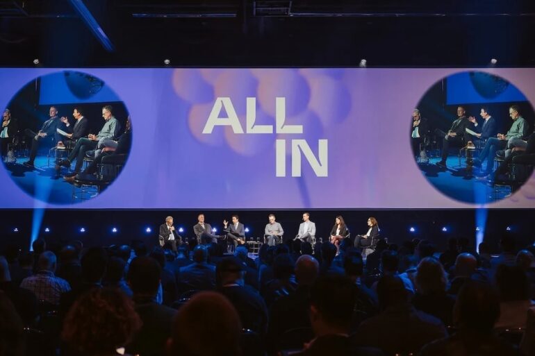 Seven people sit in a row onstage in front of a large crowd