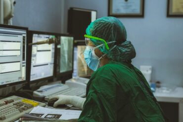 A person in green scrubs and a face mask sits at a computer terminal