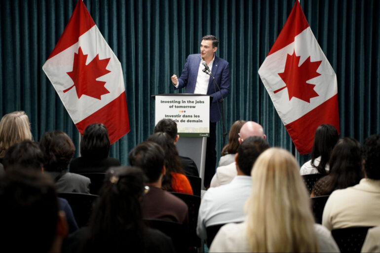 Evan Solomon speaks onstage standing at a podium between two Canadian flags