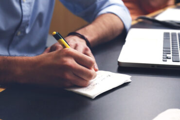 Hands writing on a notepad next to a laptop