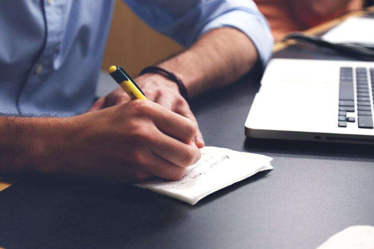 Hands writing on a notepad next to a laptop