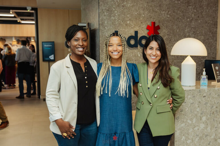 Three women stand smiling in front of a BDC sign.