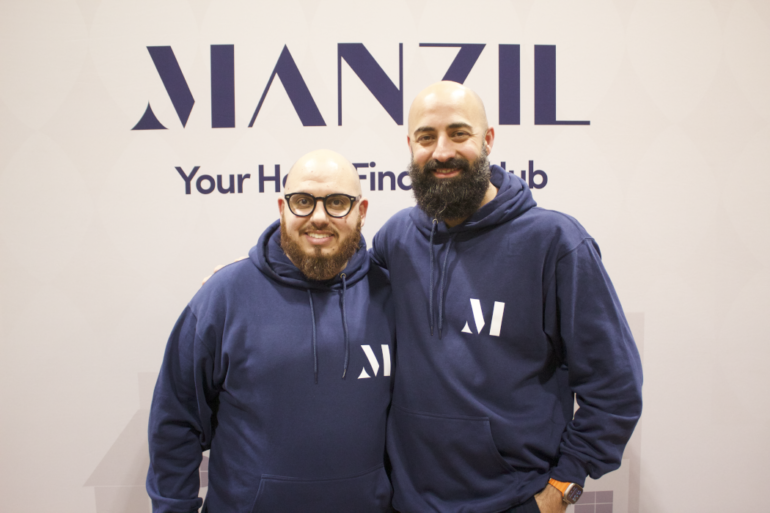 Two men stand in front of a Manzil sign with their arms on each other's shoulders.